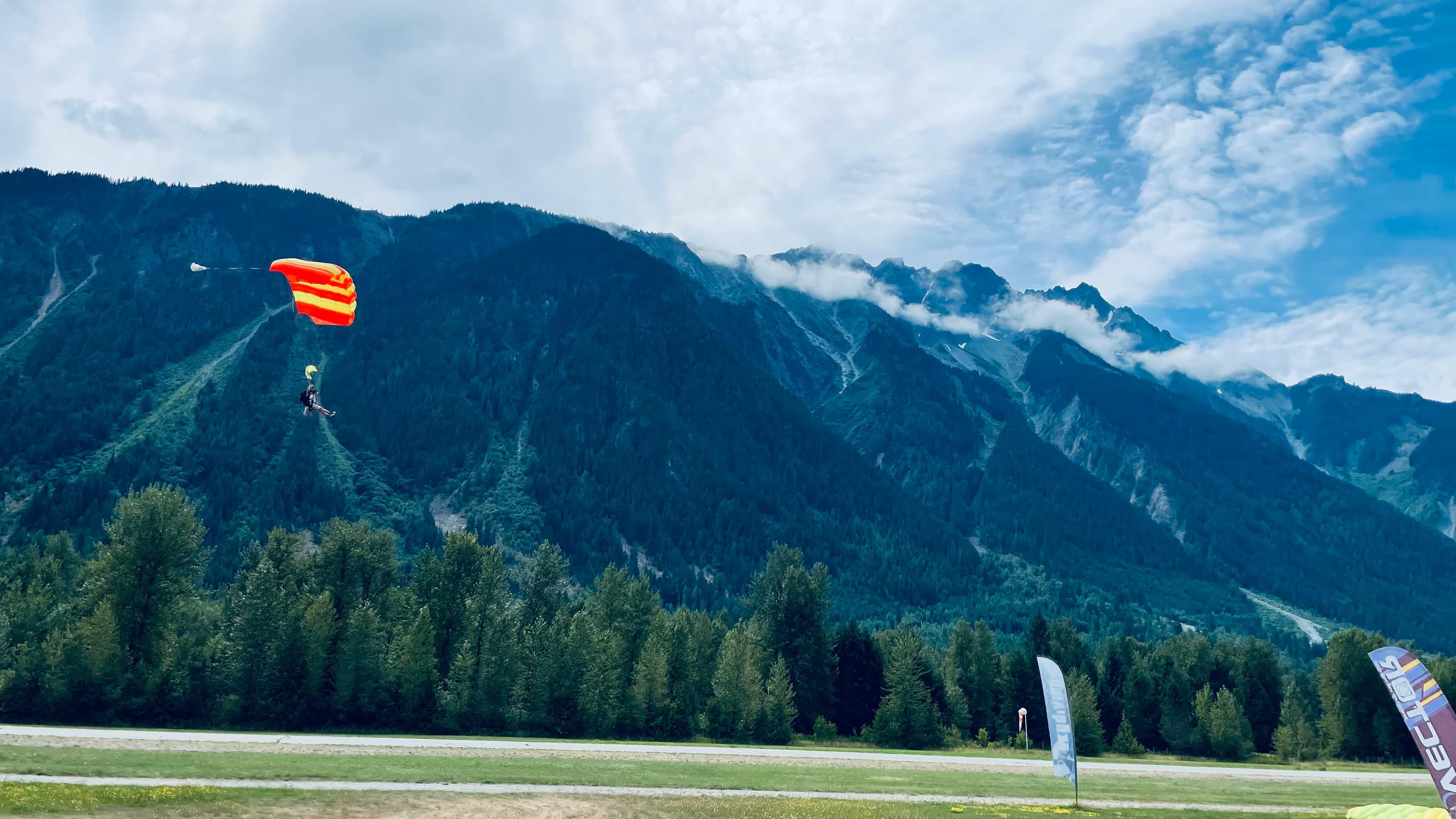 Tandem skydivers under an orange parachute coming in to land against forested mountains